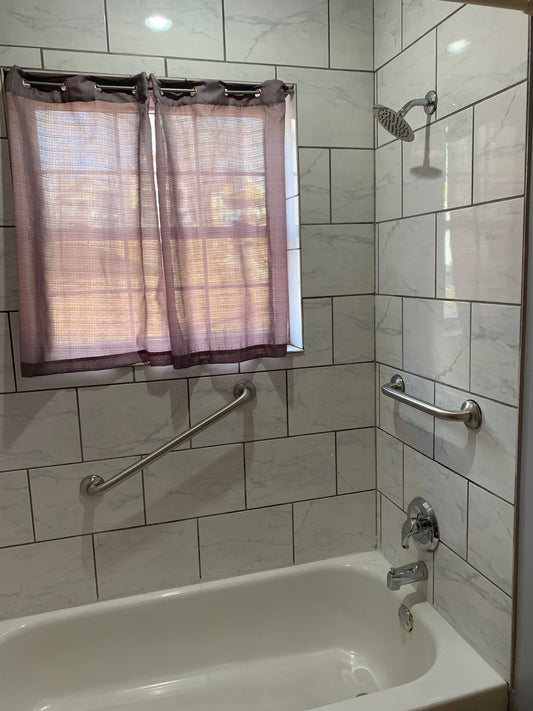 A bathroom with grab bars installed around the tub and shower area, featuring a white tub, gray tiles, and a shower curtain from AskSAMIE