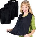 Woman seated at a table wearing a black waterproof adult bib, with a stack of folded black bibs beside her showing the 3 pack.