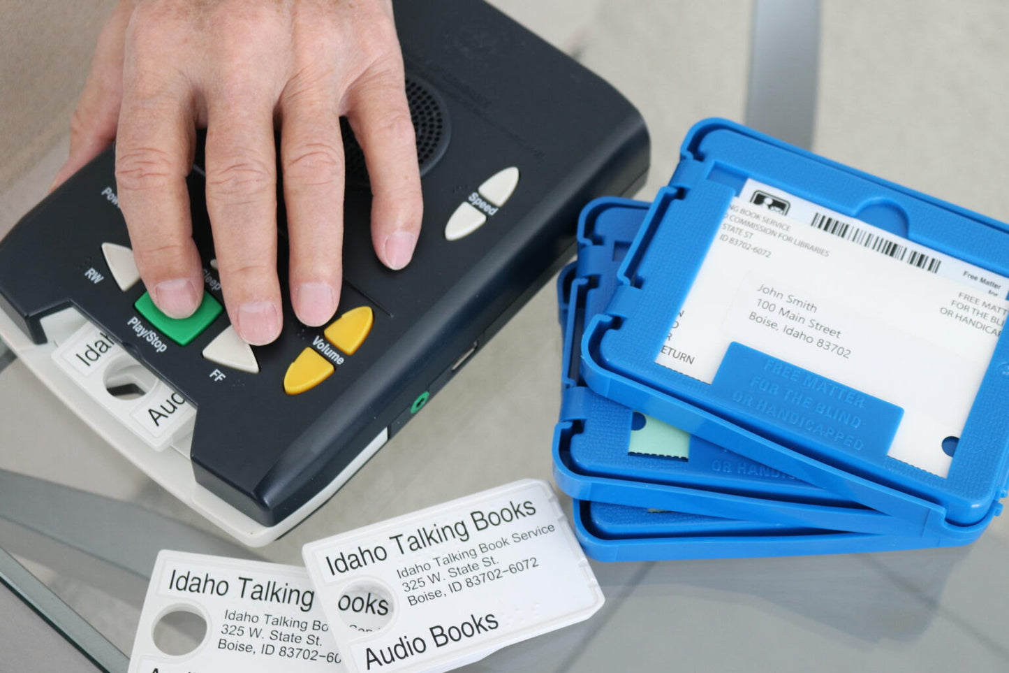 A person's hand using a blue and black audiobook player with a control panel, alongside a label that specifies the service is for 'Idaho Talking Books'. from AskSAMIE
