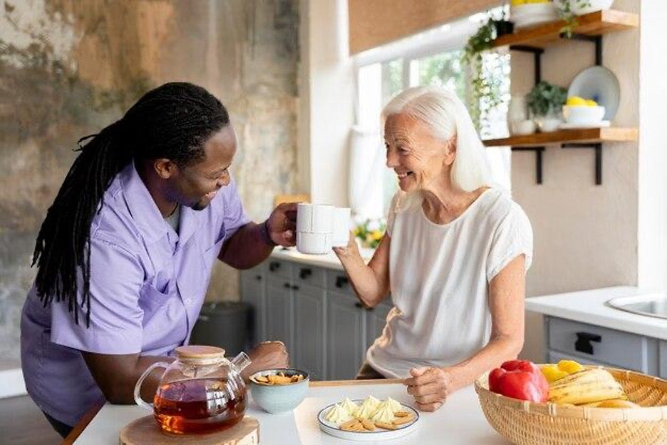 black man with long locs having coffee with an older white womn he is caring for. AskSAMIE.com the place for education for caregivers on adaptive equipment and all things aging in place.