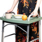 A woman walks with a folding walker carrying a coffee mug, a bowl of cereal and an orange on the green walker tray, keeping both hands on the walker handles.