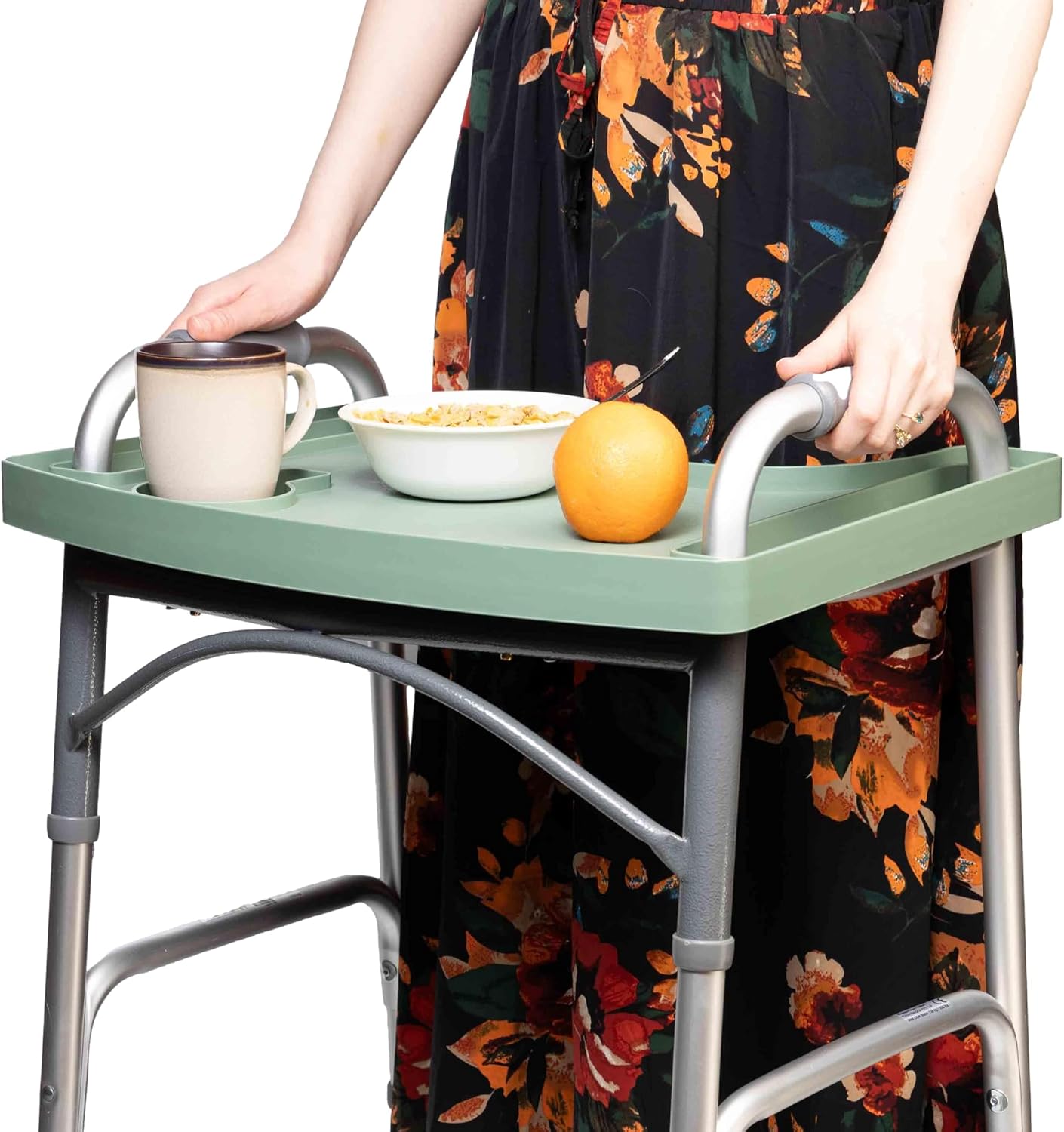 A woman walks with a folding walker carrying a coffee mug, a bowl of cereal and an orange on the green walker tray, keeping both hands on the walker handles.