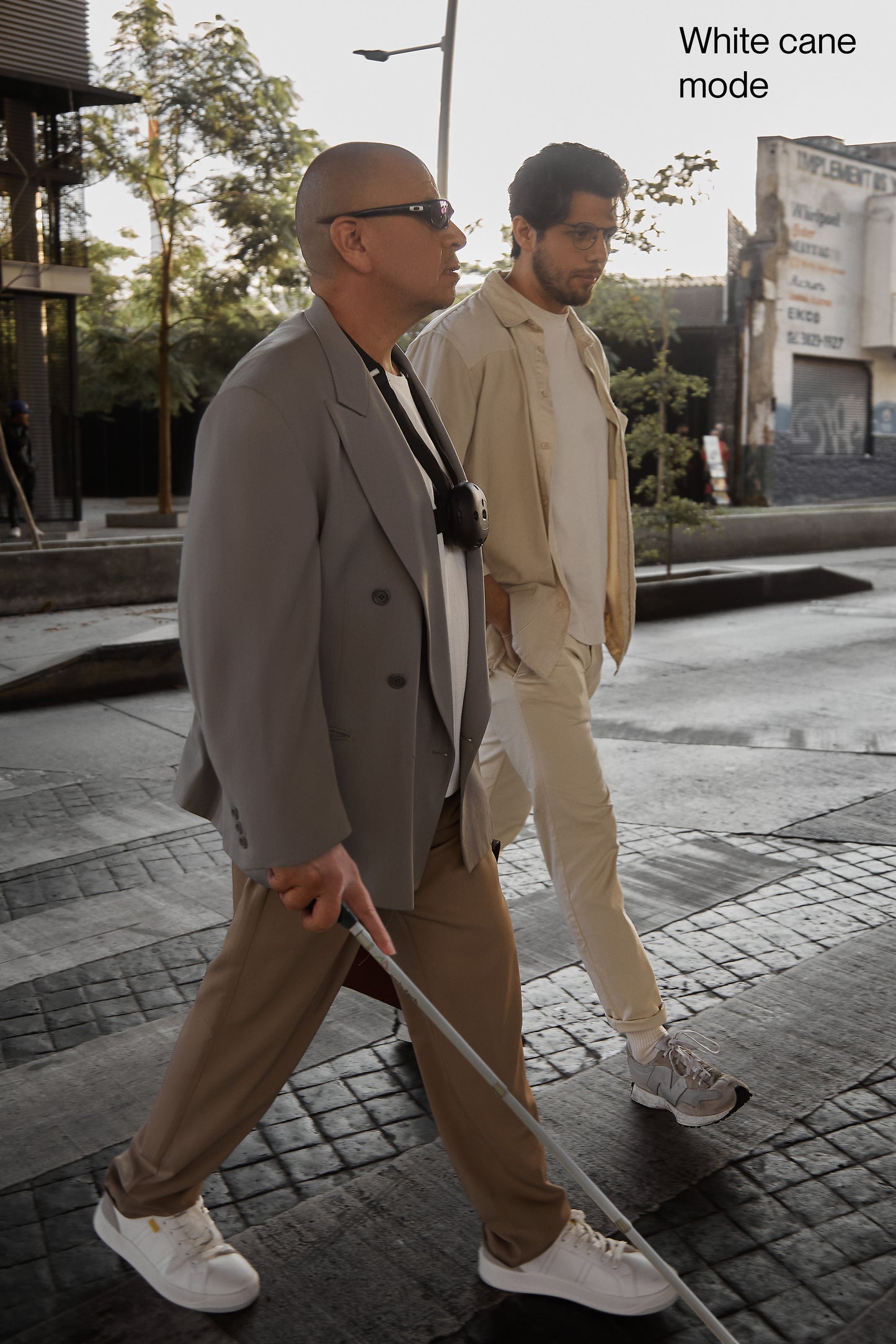 The image shows 2 men walking across a cross walk, one with glasses,  Ara and a white cane. The other man is walking along side him.