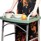 A woman walks with a folding walker carrying a coffee mug, a bowl of cereal and an orange on the green walker tray, keeping both hands on the walker handles.